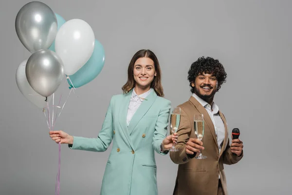 Smiling multiethnic event hosts in formal wear holding festive balloons, microphone and glasses champagne while looking at camera during party isolated on grey — Stock Photo