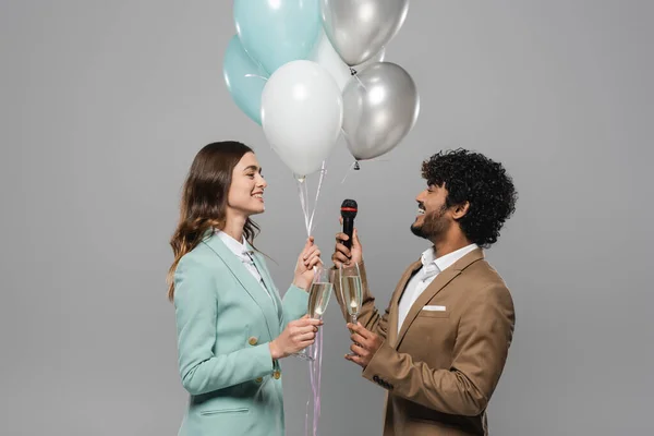 Carefree interracial event hosts in suits holding festive balloons, microphone and glasses of champagne during party while standing together isolated on grey — Stock Photo
