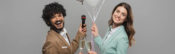 Des hôtes interraciaux positifs en costumes tenant des ballons festifs, un microphone et des verres de champagne tout en restant ensemble isolés sur une bannière grise — Photo de stock