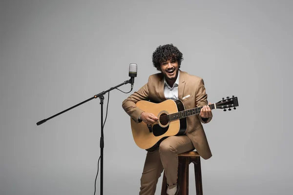 Веселый индийский организатор мероприятия в формальной одежде играет на акустической гитаре возле микрофона, сидя на стуле и глядя на камеру, изолированную на сером — стоковое фото