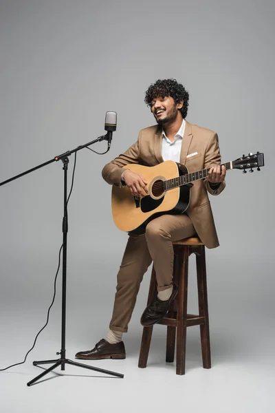 Anfitrión indio sonriente en ropa formal tocando la guitarra acústica y cantando en el micrófono en pie mientras está sentado en la silla sobre fondo gris - foto de stock