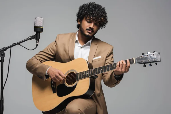 Young and curly indian host of event playing acoustic guitar near microphone on stand while sitting on chair during performance isolated on grey — Stock Photo