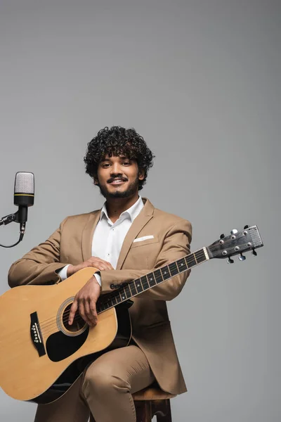 Молодой улыбающийся индиец в формальной одежде держит акустическую гитару и смотрит в камеру, сидя на стуле рядом с микрофоном, изолированным на сером — стоковое фото