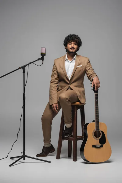 Полная длина молодого индийского организатора мероприятия в формальной одежде, держащего акустическую гитару возле микрофона, сидящего на стуле и смотрящего на камеру на сером фоне — стоковое фото
