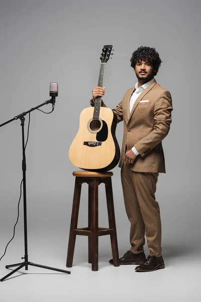 Полная длина индийского ведущего мероприятия в формальной одежде, держащего акустическую гитару возле стула и микрофона, глядя на камеру и стоя на сером фоне — стоковое фото