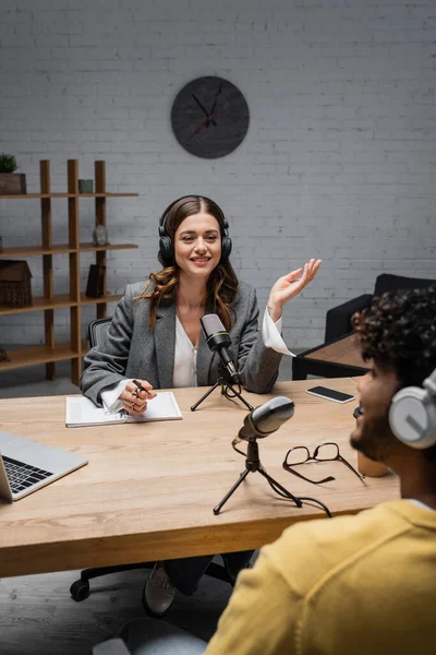 Smiling podcast host in wireless headphones talking to blurred indian guest near notebook, pen, glasses, microphones and devices with copy space during stream in studio — Stock Photo