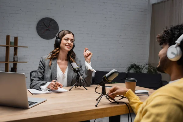 Positive female podcast host in wireless headphones talking to blurred indian guest while sitting with pen near notebook, devices, microphone and coffee to go during stream in studio — Stock Photo