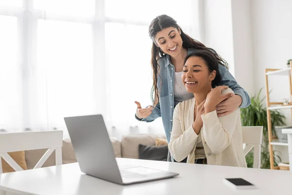 Glückliche lesbische Frau zeigt während Videoanruf auf multirassische Freundin in der Nähe von Laptop — Stockfoto