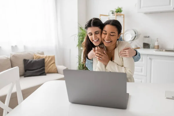 Glückliche lesbische Frau umarmt multirassische Freundin mit Verlobungsring am Finger in der Nähe von Laptop während Videoanruf — Stockfoto