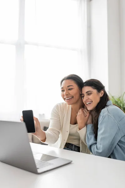 Glückliche multirassische lesbische Frau zeigt Smartphone während Videoanruf, während sie neben Freundin sitzt — Stockfoto
