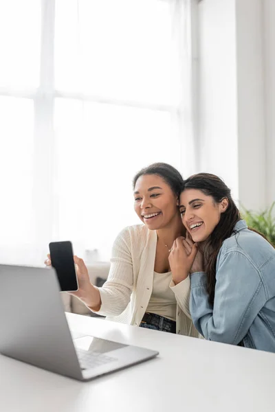 Feliz multirracial lesbiana mujer mostrando smartphone durante videollamada mientras sentado al lado de novia - foto de stock