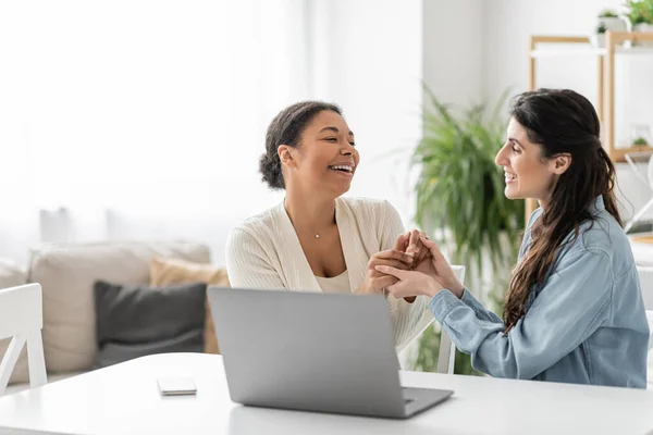 Cheerful interracial lesbian couple holding hands near gadgets on table — Stock Photo