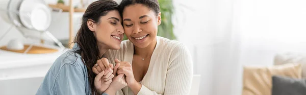 Pleased interracial lesbian couple with closed eyes holding hands at home, banner — Stock Photo