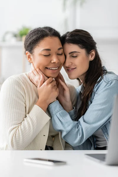 Cheerful interracial lesbian couple with closed eyes hugging near devices on blurred foreground — Stock Photo