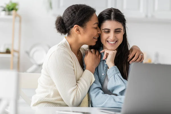 Positivo interracial lesbiana pareja con cerrado ojos abrazando cerca de dispositivos en borrosa primer plano - foto de stock