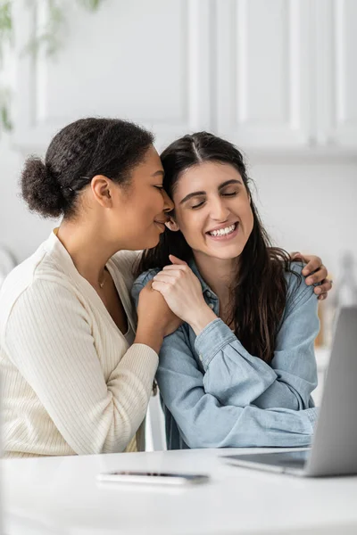 Joyful interracial lesbian couple with closed eyes hugging near devices on blurred foreground — Stock Photo