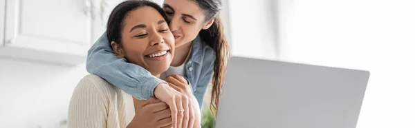 Joyeux couple lesbienne multiethnique montrant bague de fiançailles pendant l'appel vidéo sur ordinateur portable, bannière — Photo de stock