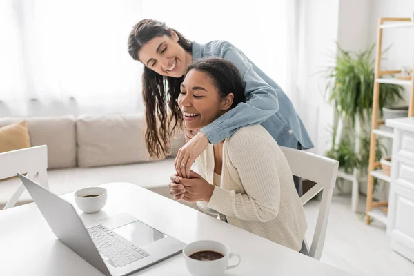 Alegre pareja lesbiana multiétnica mostrando anillo de compromiso durante la videollamada en casa - foto de stock