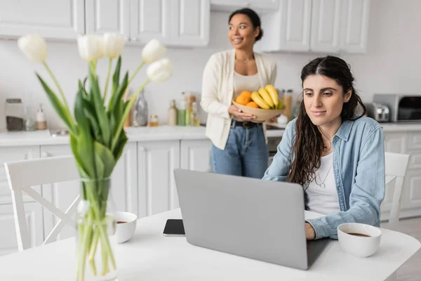 Alegre lesbiana mujer usando laptop cerca multiracial novia con bowl de frutas en borrosa fondo - foto de stock
