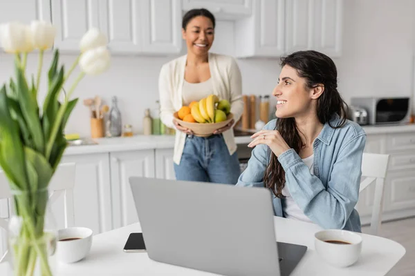 Felice donna lesbica utilizzando laptop vicino fidanzata multirazziale con ciotola di frutta su sfondo sfocato — Foto stock