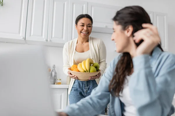 Счастливая многорасовая женщина держит миску с фруктами и глядя на подругу на размытом переднем плане — стоковое фото