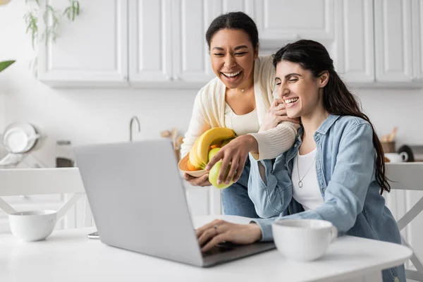 Freudige multirassische Frau hält Schale mit Früchten in der Nähe ihrer Freundin, die zu Hause am Laptop arbeitet — Stockfoto