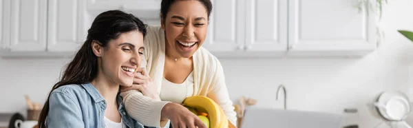 Joyeuse femme multiraciale tenant bol avec des fruits près de petite amie à la maison, bannière — Photo de stock