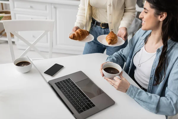 Donna che tiene piatti con croissant al forno vicino a ragazza allegra che lavora su computer portatile da casa — Foto stock