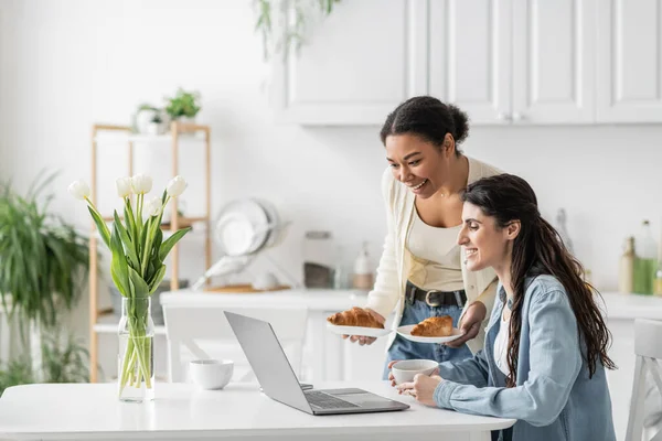 Счастливая многорасовая женщина держит тарелки с круассанами рядом с подругой работает на ноутбуке из дома — стоковое фото