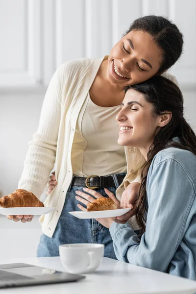 Glückliche und lesbische multirassische Frau, die Teller mit Croissants hält, während sie sich auf Freundin stützt — Stockfoto