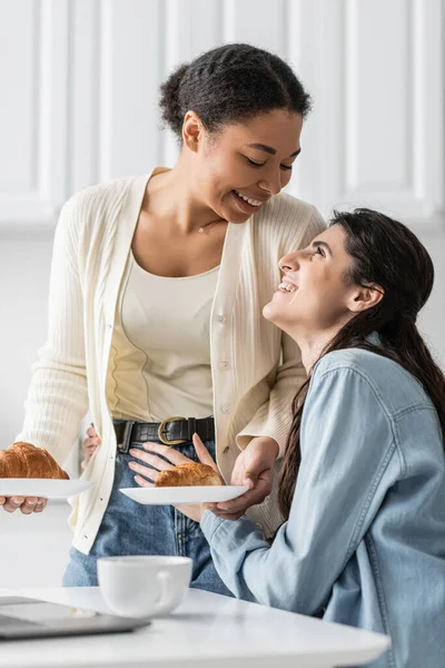 Веселая и лесбийская многорасовая женщина, держащая тарелки с круассанами, глядя на счастливую девушку — стоковое фото