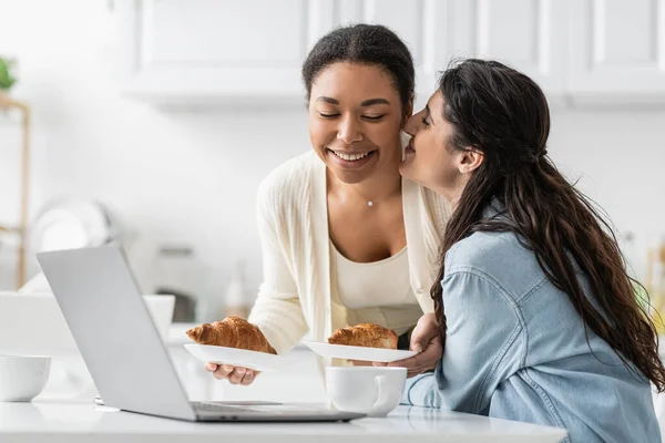 Glückliche lesbische Frau küsst Wange einer fröhlichen multirassischen Freundin mit Croissants — Stockfoto