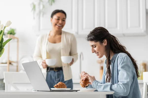 Happy lesbian woman eating croissant near laptop and multiracial girlfriend with cups on blurred background — Stock Photo