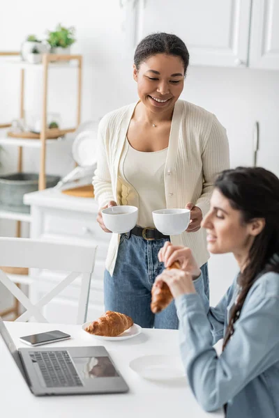 Heureuse femme multiraciale tenant tasses de café près de petite amie et gadgets sur la table — Photo de stock