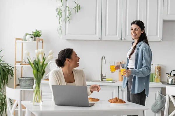 Happy lesbian woman holding glasses with orange juice near multiracial girlfriend working on laptop — Stock Photo