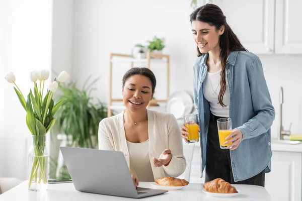 Mulher lésbica alegre segurando óculos com suco de laranja perto de namorada multirracial trabalhando no laptop — Fotografia de Stock