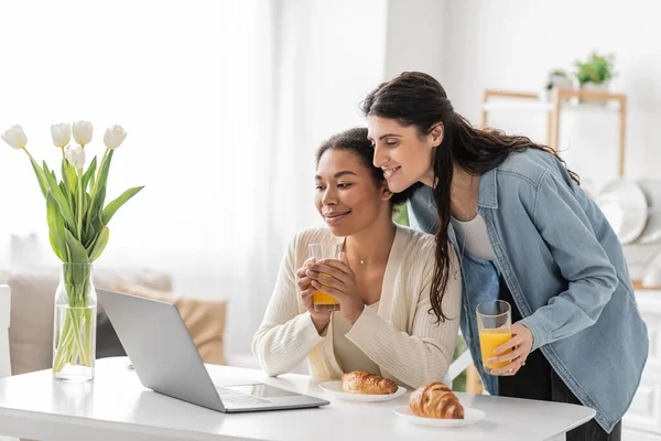 Щаслива міжрасова лесбіянка пара дивиться на ноутбук, тримаючи окуляри апельсинового соку — стокове фото