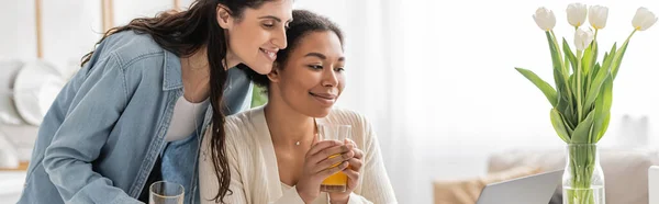 Happy interracial lesbian couple holding glasses of orange juice near tulips, banner — Stock Photo