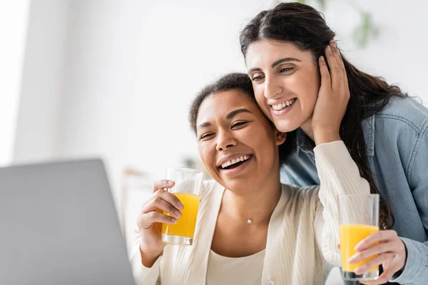 Fröhliches lesbisches Paar, das auf den Laptop schaut, während es Orangensaft in der Hand hält — Stockfoto
