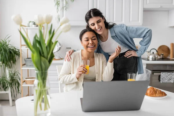 Cheerful multiracial lesbian couple looking at laptop while having breakfast in kitchen — Stock Photo