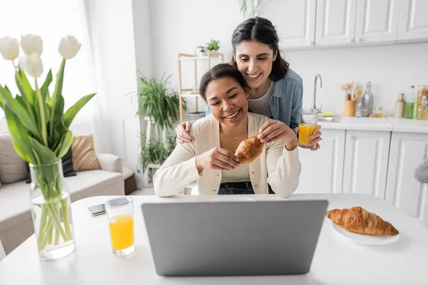 Happy multiracial lesbian couple looking at laptop while having breakfast in kitchen — Stock Photo