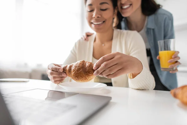 Glückliche multirassische Frau hält Croissant in der Nähe von lesbischem Partner und Laptop — Stockfoto