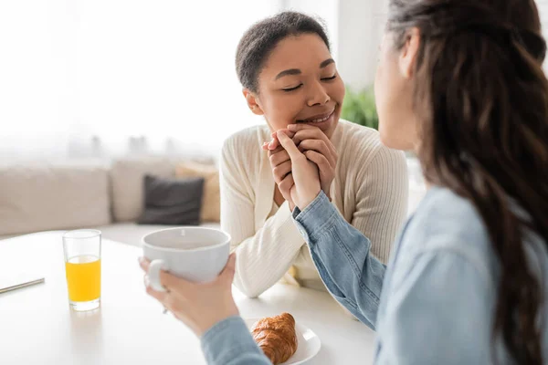 Femme lesbienne multiraciale avec les yeux fermés souriant tout en tenant la main du partenaire pendant le petit déjeuner — Photo de stock