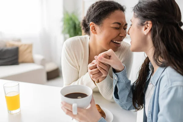 Multiracial lesbian woman with closed eyes laughing while holding hand of partner during breakfast — Stock Photo