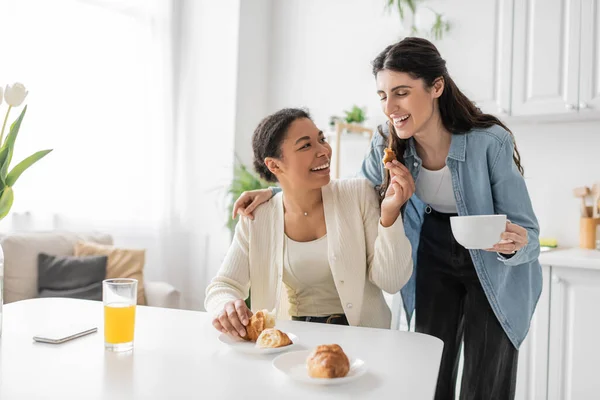 Многорасовая лесбиянка с круассаном возле подружки во время завтрака на кухне — стоковое фото