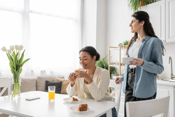 Feliz interracial lésbicas mulheres sorrindo enquanto desfruta de café da manhã na cozinha — Fotografia de Stock