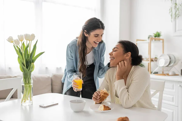 Glückliche lesbische Frau hält ein Glas Orangensaft in der Nähe ihrer multirassischen Freundin in der Küche — Stockfoto