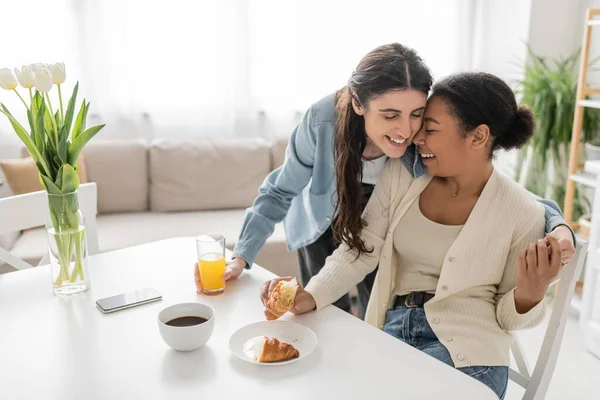 Mulher lésbica feliz segurando vidro de suco de laranja e abraçando namorada multirracial durante o café da manhã na cozinha — Fotografia de Stock