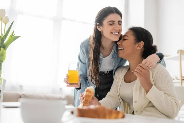 Mulher lésbica alegre segurando vidro de suco de laranja e abraçando namorada multirracial durante o café da manhã na cozinha — Fotografia de Stock