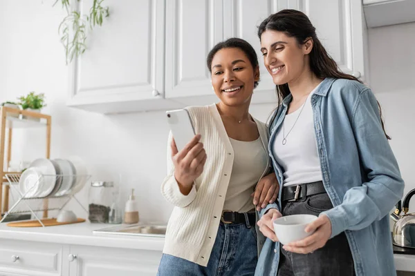 Happy multiracial woman holding smartphone near lesbian partner with cup of coffee — Stock Photo